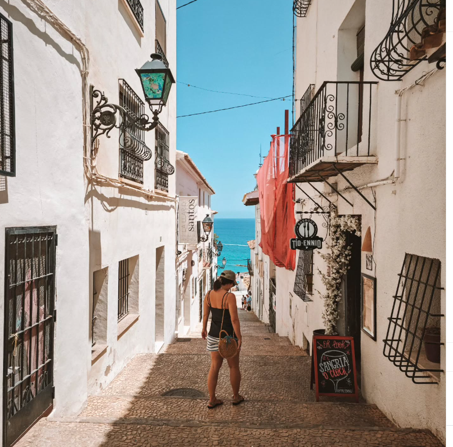 Luana caminhando por uma ruela branca de Altea, com o mar Mediterrâneo ao fundo