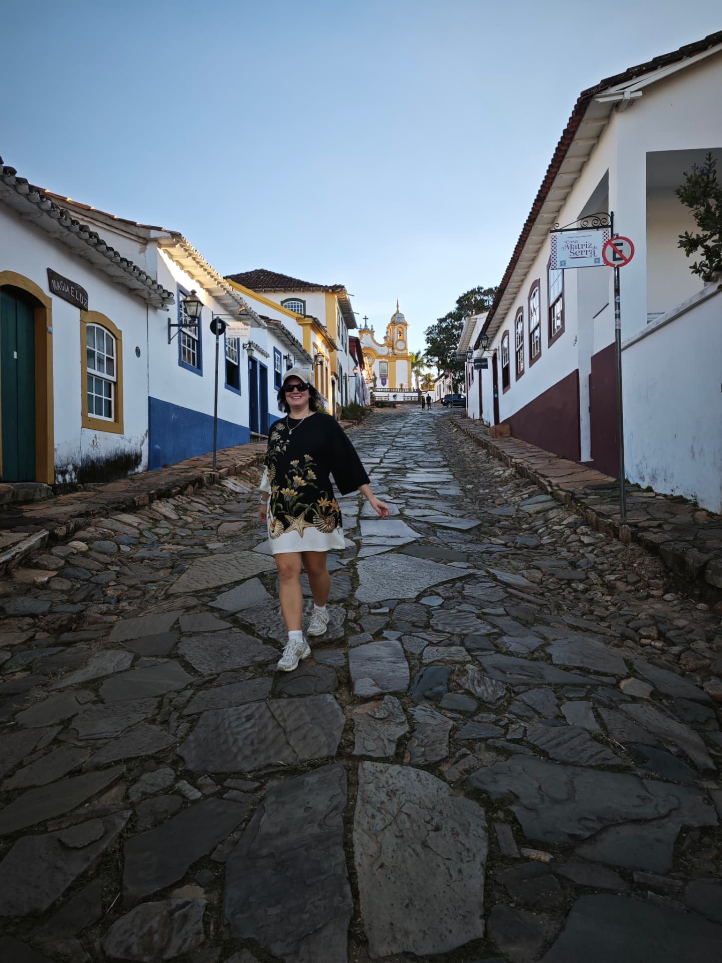 Luana caminhando por uma rua de pedras de Tiradentes, com a igreja ao fundo